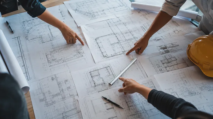 Top-down view of a meeting table covered with multiple unrolled large-format construction drawings, scale ruler and pen on top, three pairs of hands pointing at details mid-coordination